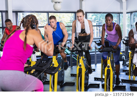 Fitness instructor leading diverse spin class participants pedaling on yellow bikes in gym studio Fitness instructor leading diverse spin class participants pedaling on yellow bikes in gym studio 135715049