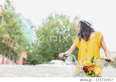Woman in twenties riding bicycle on cobblestones wearing sunhat carrying flower basket, copy space Woman in twenties riding bicycle on cobblestones wearing sunhat carrying flower basket, copy space 135715057