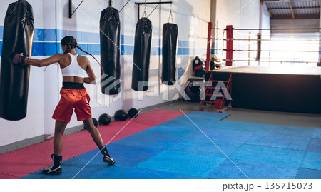 Female athlete wearing boxing shorts and gloves practicing boxing with heavy bags in boxing gym 135715073