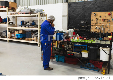 Man wearing coveralls hairnet talking on smartphone while sorting labeled bins at workshop bench Man wearing coveralls hairnet talking on smartphone while sorting labeled bins at workshop bench 135715081