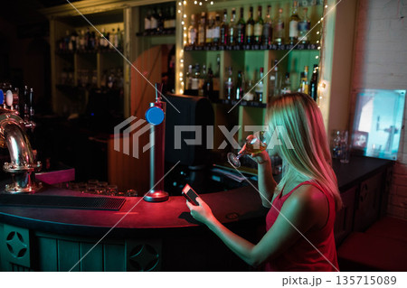 Woman standing at bar under red and green lights holding smartphone and wine glass, copy space 135715089
