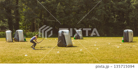 Teenage female archer crouching on field, aiming recurve bow amid bunkers, marker cones, copy space 135715094