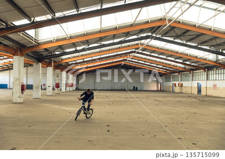 Man performing BMX bicycle stunt inside industrial warehouse wearing dark t-shirt and pants 135715099