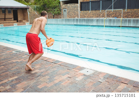 Male lifeguard holding orange rescue float leaning in on brick deck at lane-marked pool, copy space Male lifeguard holding orange rescue float leaning in on brick deck at lane-marked pool, copy space 135715101