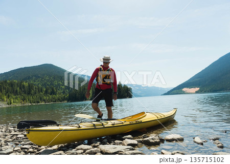 Senior man balancing on yellow kayak at mountain lake shore, holding paddle, wearing life vest, hat 135715102