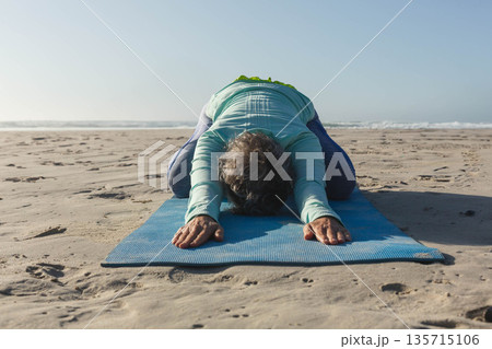 Senior woman practicing yoga on blue mat on sandy beach in teal-purple gear with green towel 135715106