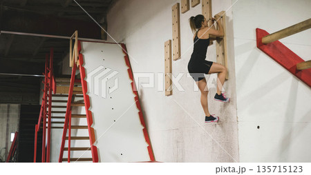 Female athlete gripping and climbing wooden pegboard wall at obstacle course facility, copy space Female athlete gripping and climbing wooden pegboard wall at obstacle course facility, copy space 135715123
