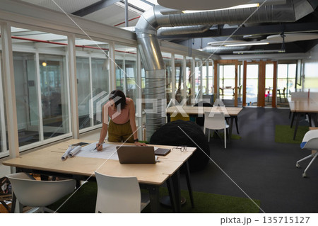 Female professional reviewing rolled blueprints at modern office desk with laptop, wearing pink top Female professional reviewing rolled blueprints at modern office desk with laptop, wearing pink top 135715127
