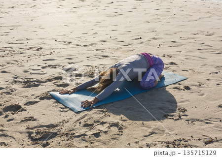 Woman kneeling into child pose on sandy beach wearing leggings using blue yoga mat in sunlight 135715129