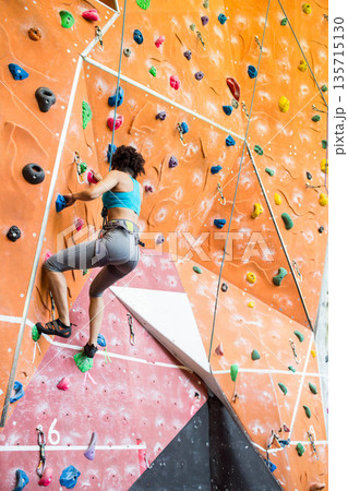 African American female climber ascending wall at gym gripping colorful holds using safety harness 135715130
