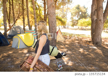 Woman holding rolled rug and sleeping mat while standing at campsite among pop-up tents, copy space 135715134