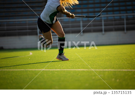 Teenage girl field hockey player sprinting across artificial turf pitch and swinging hockey stick Teenage girl field hockey player sprinting across artificial turf pitch and swinging hockey stick 135715135