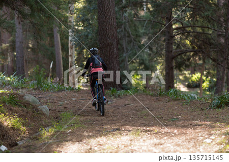 Female cyclist riding narrow dirt trail in dense forest on mountain bike with helmet, backpack Female cyclist riding narrow dirt trail in dense forest on mountain bike with helmet, backpack 135715145