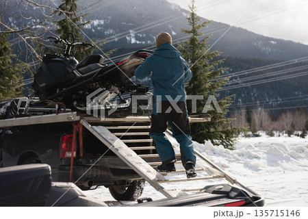 Mid adult man in winter jacket guiding snowmobile up ramp into pickup truck at forest trail 135715146