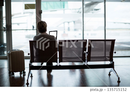 Middle-aged businesswoman sitting on bench at airport gate using tablet with suitcase, copy space Middle-aged businesswoman sitting on bench at airport gate using tablet with suitcase, copy space 135715152