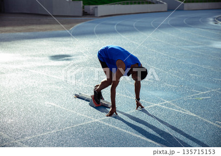 African American adult male athlete aligning in sprint start stance on blue track with metal blocks African American adult male athlete aligning in sprint start stance on blue track with metal blocks 135715153