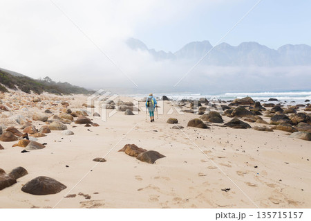 Woman walking on sandy beach wearing wide-brim hat and carrying backpack using trekking poles 135715157