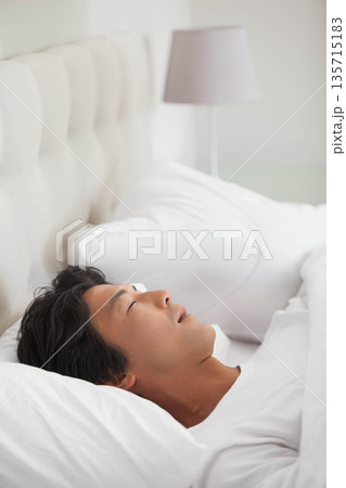 Asian man lying on bed in bedroom with upholstered headboard, white duvet and gray lamp Asian man lying on bed in bedroom with upholstered headboard, white duvet and gray lamp 135715183