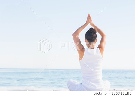 Woman wearing white outfit raising arms practicing yoga on sandy shore facing calm sea horizon 135715190