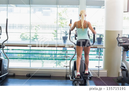 Female gymgoer working out on elliptical in cardio area by windows overlooking pool and plants Female gymgoer working out on elliptical in cardio area by windows overlooking pool and plants 135715191