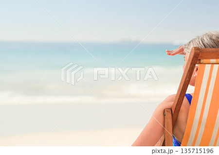 Senior woman sitting in orange-striped beach chair at sandy shore shading eyes from sun, copy space Senior woman sitting in orange-striped beach chair at sandy shore shading eyes from sun, copy space 135715196