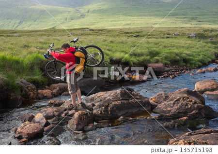 Male wearing red windbreaker and yellow backpack crossing stream with mountain bike on moorland 135715198