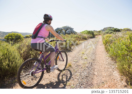 Adult woman cycling along dirt trail on purple mountain bike wearing helmet and hydration backpack Adult woman cycling along dirt trail on purple mountain bike wearing helmet and hydration backpack 135715201