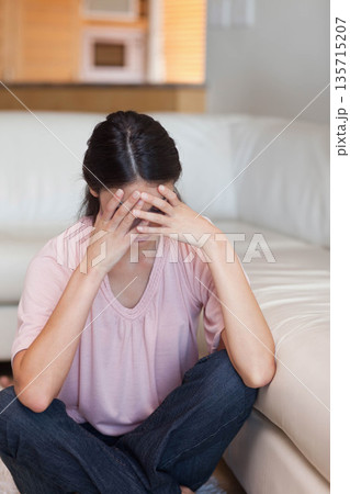 Woman wearing jeans sitting against white couch in living room under window blinds near wooden sill 135715207