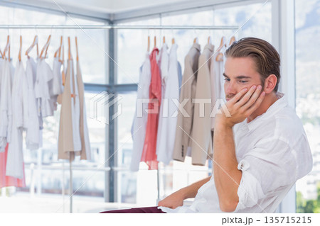 Man wearing white shirt sitting on stool in clothing showroom examining shirts on rack, copy space 135715215