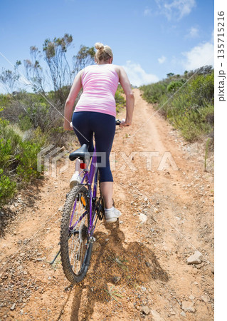 Woman riding purple bike with white water bottle, pedaling uphill on hillside trail in sportswear Woman riding purple bike with white water bottle, pedaling uphill on hillside trail in sportswear 135715216