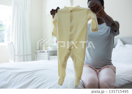 African American woman holding pale yellow footed baby onesie and sitting on bed edge in bedroom African American woman holding pale yellow footed baby onesie and sitting on bed edge in bedroom 135715495