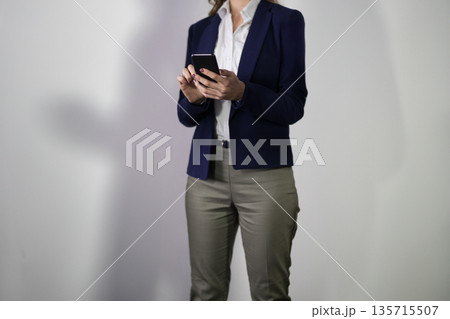 Businesswoman in blazer and trousers scrolling through messages on smartphone against gray backdrop Businesswoman in blazer and trousers scrolling through messages on smartphone against gray backdrop 135715507
