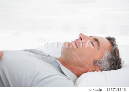 Middle-aged man resting on white bedding and pillow in bedroom wearing light grey button-up shirt Middle-aged man resting on white bedding and pillow in bedroom wearing light grey button-up shirt 135715546
