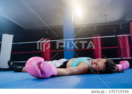 Female boxer lying in boxing ring wearing pink gloves, turquoise top among punching bags and ropes 135715548