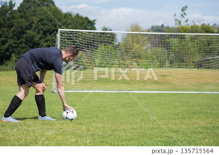 Male soccer player in black uniform placing ball on grass pitch beside goal frame, copy space 135715564