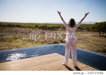 Woman standing with arms raised on wooden deck at infinity pool overlooking distant hills 135715566
