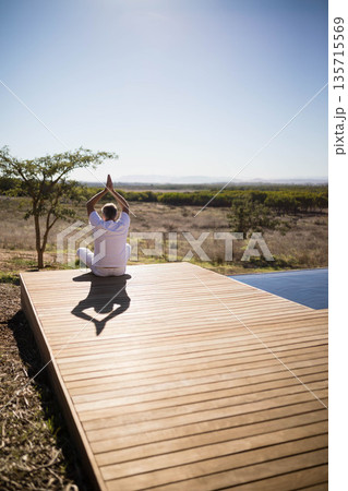 Man sitting in lotus position on wooden deck by small tree overlooking grassland and pool edge Man sitting in lotus position on wooden deck by small tree overlooking grassland and pool edge 135715569