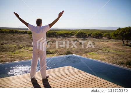 Middle-aged male stretching arms out on wooden pool deck by infinity pool overlooking scrub plain Middle-aged male stretching arms out on wooden pool deck by infinity pool overlooking scrub plain 135715570