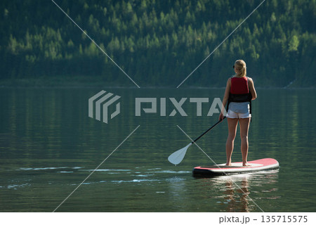 Female paddleboarder paddling on red paddleboard in lake wearing red vest, shorts, copy space Female paddleboarder paddling on red paddleboard in lake wearing red vest, shorts, copy space 135715575