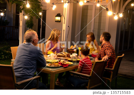 Multi generation family chatting around wicker dining table on backyard patio under string lights Multi generation family chatting around wicker dining table on backyard patio under string lights 135715583