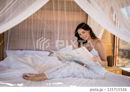 woman sitting cross-legged on bed beneath sheer canopy reading paper map in resort bungalow bedroom 135715588