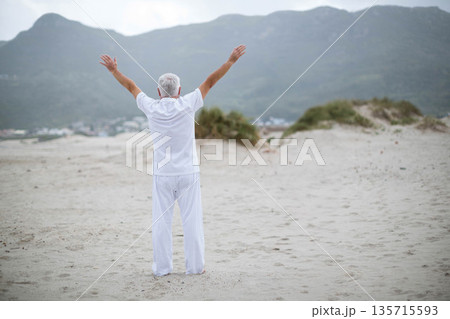 Senior man standing on wide sandy shoreline near dunes facing coastal town under overcast sky 135715593