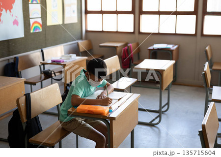 Child boy wearing green shirt and mask writing on paper in classroom with orange pencil case Child boy wearing green shirt and mask writing on paper in classroom with orange pencil case 135715604