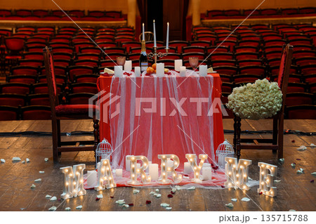 Romantic proposal setup in grand theater with red velvet seats, candlelit table, and illuminated "MARRY ME" letters Romantic proposal setup in grand theater with red velvet seats, candlelit table, and illuminated "MARRY ME" letters 135715788