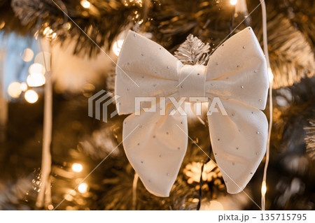 Elegant White Bow Ornament Adorned with Sparkling Rhinestones Hanging on a Festive Christmas Tree with Warm Lights Elegant White Bow Ornament Adorned with Sparkling Rhinestones Hanging on a Festive Christmas Tree with Warm Lights 135715795