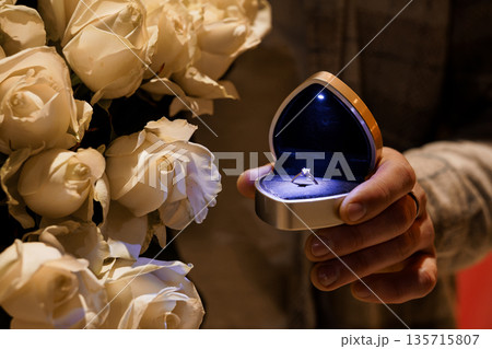 Heart-shaped ring box with diamond engagement ring illuminated by blue light beside white roses bouquet Heart-shaped ring box with diamond engagement ring illuminated by blue light beside white roses bouquet 135715807