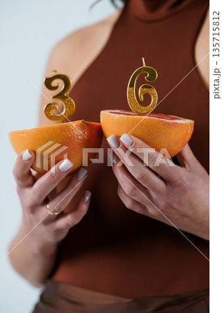 Woman holding grapefruit halves with golden number candles 3 and 6 for birthday celebration in warm brown tones 135715812