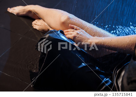 Wet woman in black lingerie lying on dark floor under blue shower rain light effect 135715841