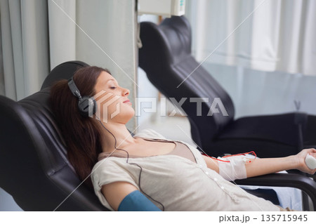 Woman reclining in clinic wearing beige top, headphones, squeezing ball, donating blood via tubing 135715945