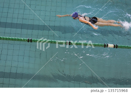 Female swimmer wearing black one-piece, purple cap, goggles, swimming past lane divider, copy space 135715968